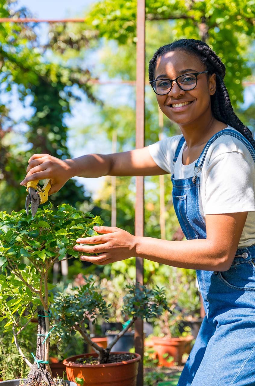 Mujer con trenzas trabajando como jardinera en un vivero, cuidando plantas y flores en un entorno natural.